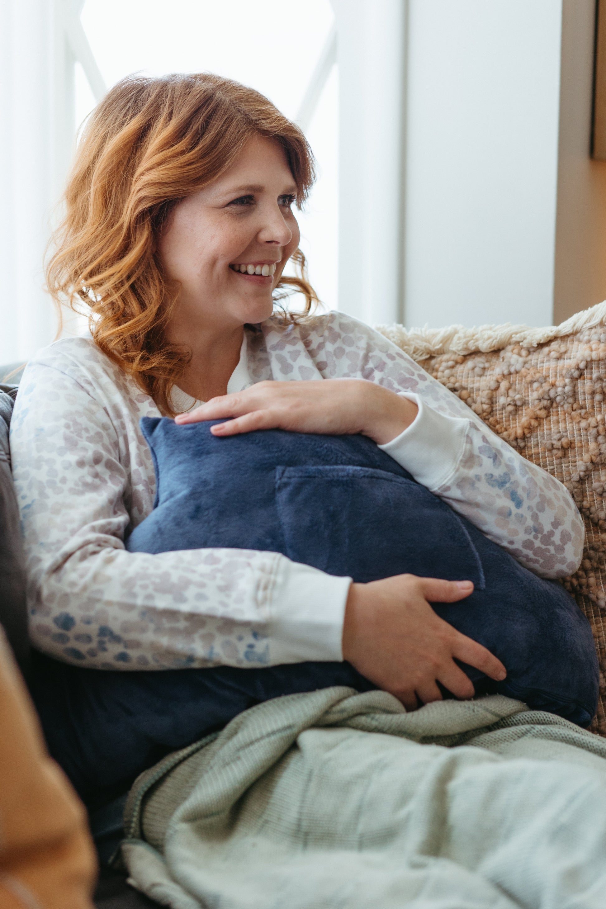 A woman smiling on the couch holding a tummy tuck recovery pillow
