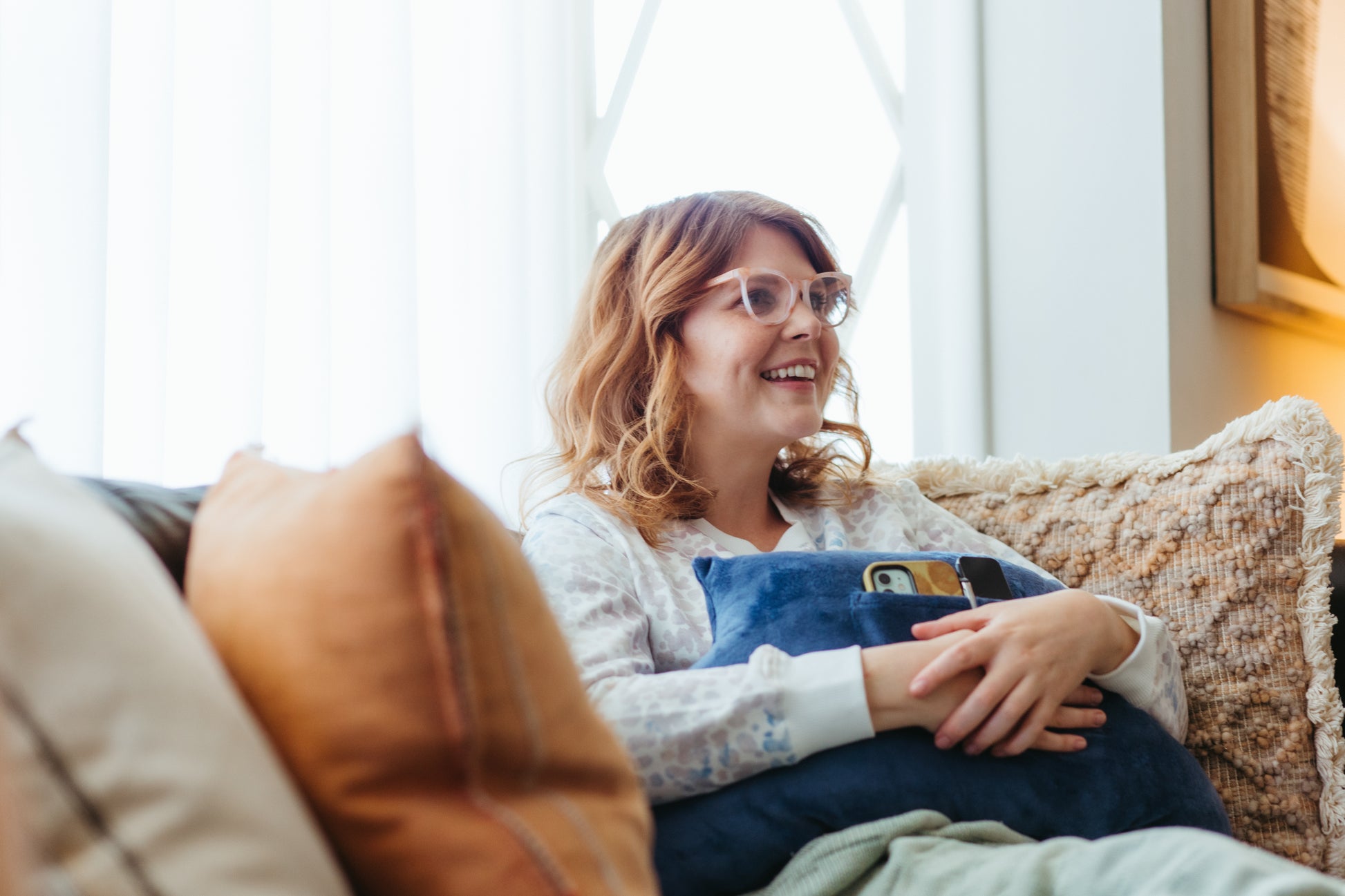 A woman holding her tummy tuck pillow on the couch with her phone in the pocket.