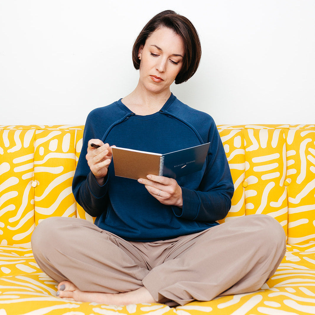 A woman writing in the Mended Wing Recovery Journal sitting on a yellow and white patterned couch